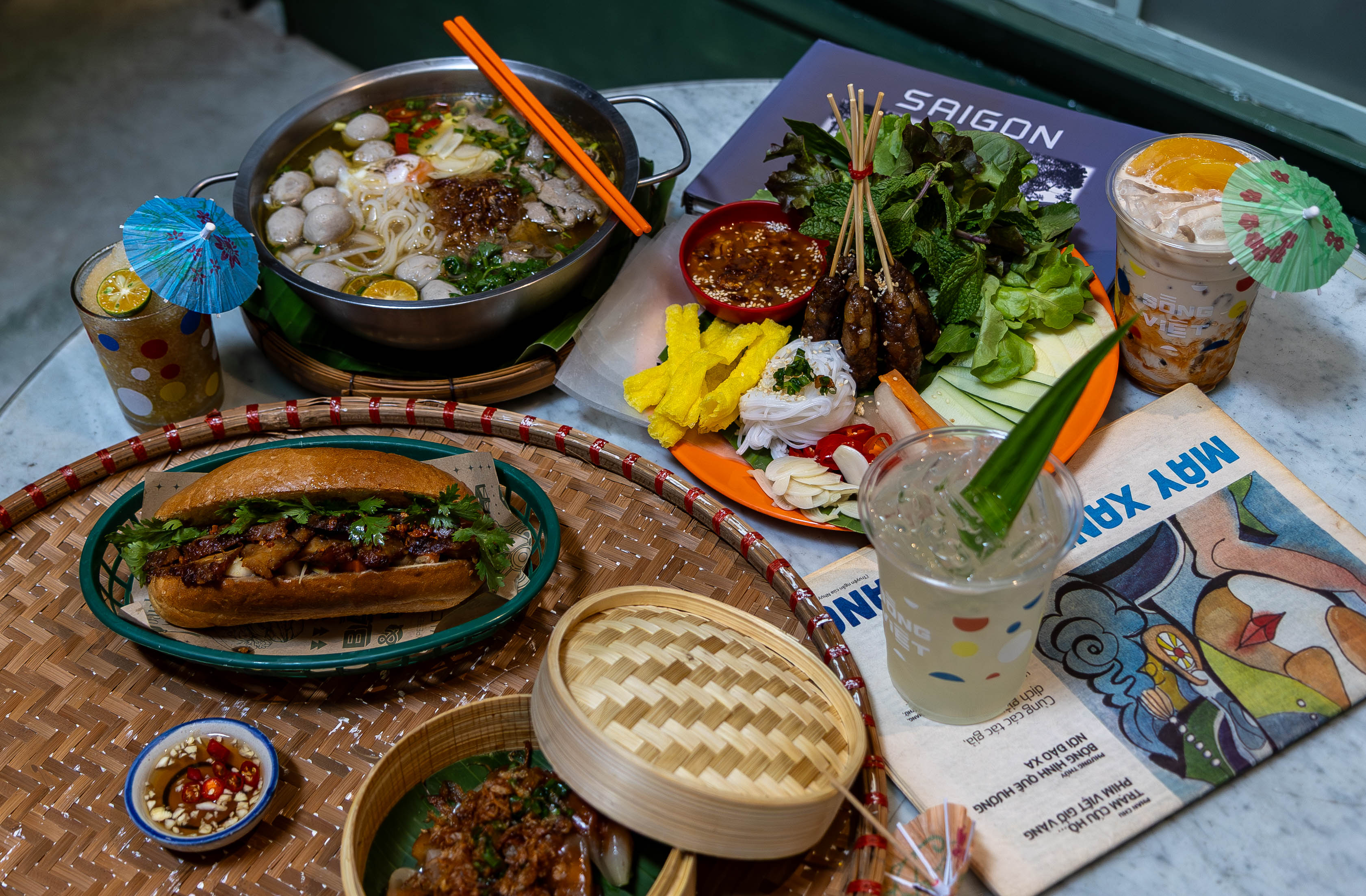 Full Vietnamese street food table at Song Viet at Song Wat — Pho, Banh Mi, Nem Nuong, bamboo steamer and drinks alongside vintage Vietnamese magazine on marble table Bangkok